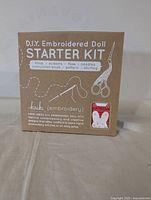 Boxed Fox D.I.Y. Embroidered Doll Starter Kit showing the front label listing contents including hoop, scissors, floss, needles, instruction book, pattern, and stuffing. Small fox embroidery is shown on packaging.