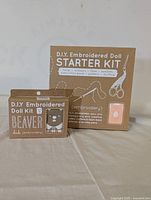 Photo showing two cardboard craft kits: A D.I.Y. Embroidered Doll Starter Kit including sewing supplies and an instruction book, and a smaller box labeled Beaver D.I.Y. Embroidered Doll Kit.
