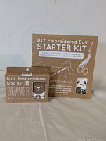 Photo of DIY Embroidered Doll Starter Kit packaging for panda and beaver dolls showing kit inclusions such as scissors, floss, needles, patterns, instruction book, and stuffing.