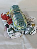 Three rolled cotton tea towels tied with twine showing varied patterns including plaid and vegetable-themed prints.