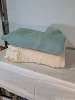 Two wool blankets stacked, teal on top, white with knitted texture and satin trim underneath