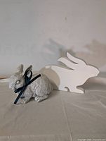 Photo showing gray acrylic bunny with a black ribbon and a white painted wooden cut-out bunny figure side by side on cloth background.
