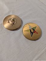 Two vintage round metal makeup compacts closed showing lids with colorful star emblem and female profile illustration.