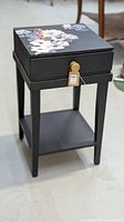 Compact black bedside table with floral decal on top, showing the front and side angles and drawer detail.