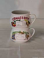 Two ceramic soup cups stacked, one labeled Tomato Soup with printed recipe and illustrations, the other labeled Onion Soup with recipe and illustrations, both white ceramic with handles.