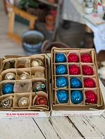 Overview of two boxes with 24 total vintage Christmas glass ornaments in red, blue, silver, white and metallic finishes, packed with cardboard dividers and tissue paper.