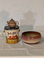 Front and side view of vintage Jack & Jill Peanut Butter tin, ceramic bowl, and teapot piggy bank placed next to each other on a cloth.