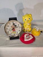 Photo showing all four vintage items on a plain surface with shadows cast behind: Mickey Mouse wristwatch, yellow dog piggy bank, red and white Mickey Mouse cap, yellow wind-up toy.