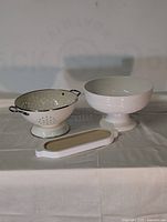 Photo of three white housewares: metal strainer colander, ceramic pedestal bowl, and wooden tray with white frame on cloth background.