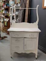 Front perspective of a vintage white-painted wooden wash stand showing three drawers, one cabinet door, and a decorative backsplash with curved vertical supports.