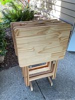 Four natural wood folding TV trays stored on accompanying wooden stand outside on concrete patio, showing wood grain and tray tops with minor blemishes.