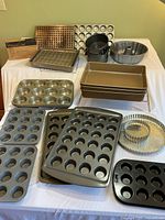 Full overview of various metal baking pans and tins including Calphalon 9x13 pans, muffin tins of different sizes, a bundt pan, tart pans, and baking racks arranged on a white surface.