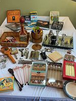 Full overview of barware items including books, cocktail stir sticks in wooden holder, metal bar tools, glass stir sticks, boxed cocktail stirrer sets and coasters on white tablecloth.