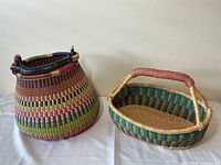 Two colorful woven baskets with leather handle covers displayed on a white cloth against a wall.
