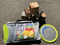 Photo shows all items together: bocce ball set in a carrying bag, yellow frisbee, and pile of large wooden dice cubes.
