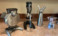 Photo showing all four items: Cuisinart waffle maker, black manual juice press, ISI stainless steel whipped cream dispenser, and Bialetti moka pot espresso maker on countertop.