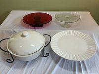 Set of cake stands and serving dish displayed on white cloth: white lidded stoneware dish on black metal stand, red glass cake stand, clear glass cake stand, and large white ceramic cake stand.