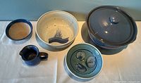 Overview of all five ceramic pieces on white tablecloth: large M. Johnson bowl, textured blue-green bowl, small blue creamer, green and blue swirl bowl, and large blue crock with lid.