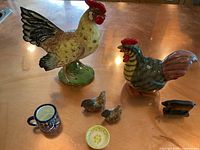 Two large ceramic chicken figurines, two small ceramic chicken figurines, colorful cup, small bowl, and miniature cast iron flat iron shown on wooden surface.