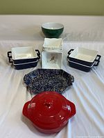 Overview photo showing all ceramic items arranged: four blue Home Essentials baking dishes stacked in pairs, green Emile Henry bowl, petite maison Wildly Delicious decorative piece, blue-white speckled baking dish, and red lidded pot.