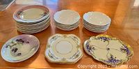 Several stacks of plates showing different designs: 11 pink and cream colored plates, 12 cream textured rim floral plates, and 1 small violet floral platter on the table.