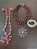 Full view of the jewelry collection including triple-strand cranberry necklace, long pink bead necklace with matching earrings, pink beaded stretchy bracelet, and large floral brooch