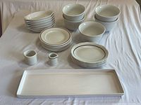 Photo showing the full dish set including stacked white bowls, small plates with rim patterns, two small creamers, and a long rectangular serving platter on a white cloth covered surface.