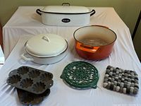 Full view of all cookware items including enameled pans, cast iron pots, and trivets on a white cloth background.