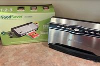 FoodSaver FM2900 vacuum sealer next to its original box, showing front and top view of the sealer unit.