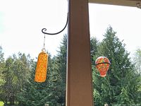 Two colorful glass hanging art pieces displayed on a patio post outdoor; one elongated orange speckled piece and one smaller rounded multi-color speckled piece