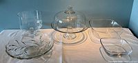 Wide shot of all glass items together on a white surface against blue wall showing the fruit bowl, two modern bowls, glass container, and cake dome.