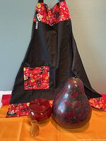 Black and red Day of the Dead themed kitty apron with patterned pocket, large dark red gourd, red wooden container, and small amber candle holder.