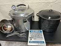 Pressure canner, enamelware pot, manual, sieves, pestle and rack posts on black surface