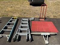 Full view of two ladders, red hand truck, and two red floor mats laid out on asphalt