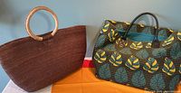 Photo showing both bags side by side to display size and style differences: brown straw bag with circular handles and patterned fabric tote with black handles.