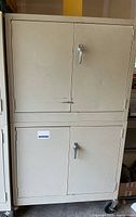 Closed view of large off-white metal cabinet with two sets of double doors and metal handles. The cabinet is on wheels.