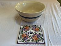 Large cream-colored stoneware bowl with two simple blue stripes around the middle paired with a colorful painted ceramic trivet featuring floral and bird designs, both items displayed on a white background.