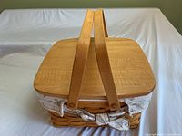 Front angled view of Longaberger woven picnic basket showing wooden lid, curved handles, and fabric liner with floral/leaf pattern tied at sides.