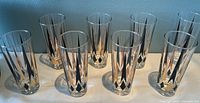 Eight tall Collins glasses with black and gold geometric designs arranged against a blue wall.