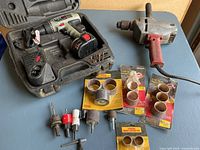 Milwaukee heavy duty corded drill, Skil 14.4V cordless drill in case with charger, drill bits, and sanding drum attachments arranged on table