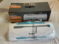 View of sealed Scotch Pro Thermal Laminator on a table in front of packaging box showing title and product image.