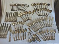 Full view of all flatware items arranged on white cloth, showing variety of knives, forks, spoons and serving utensils with ornate handles and tarnish.