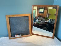 Front view of small square slate chalkboard with wood frame and rectangular wood framed mirror beside it on table.