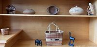Shelf display showing a dark wood carved elephant, a smaller woven basket, three flat and round woven baskets of different sizes with lids, and a wood carved light-colored eagle figure in flight on base.