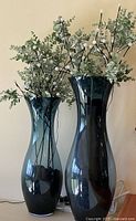 Full view of two tall blue glass vases containing faux eucalyptus plants with LED lights, showing relative size and shape.