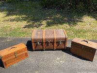 Photo of three storage items placed outside on pavement with grass background: large Unica trunk in center and two wooden boxes on sides.
