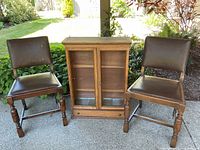 Pair of vintage upholstered chairs flanking antique wooden cabinet with glass doors on patio.