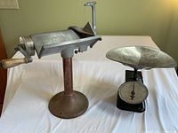 Front view of the Pelouze scale with round pan and dial indicator next to Schor cheese slicer with hand crank and metal base stand on white tablecloth.