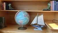 Vintage world globe, model sailboat, and bronze patina bookend on wooden shelf.