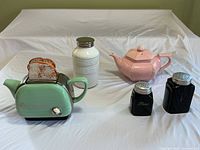 Full lot showing all six items arranged on a white cloth: one white ceramic jar, pink teapot, jade green tea toaster teapot, black shakers, and toaster-shaped measuring cup stack with bread slices.